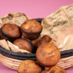 Close-up of a wicker basket filled with freshly baked cinnamon muffins, with decorative paper and dried foliage, on a pink background
