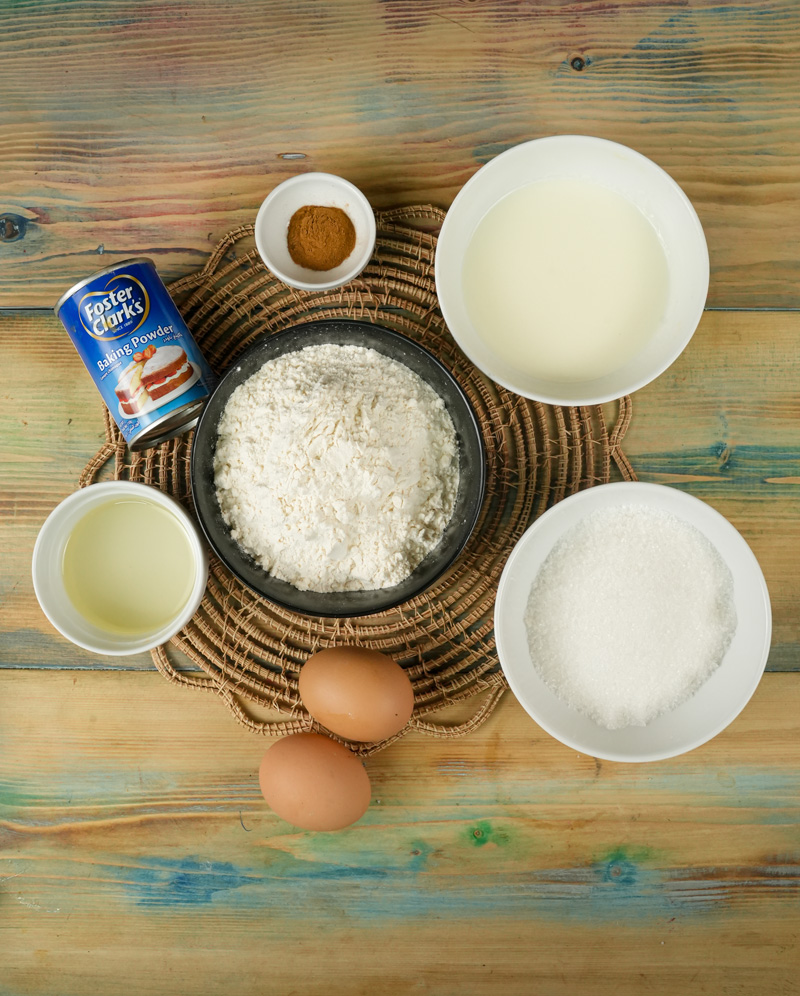 Ingredients for cinnamon muffins including flour, sugar, eggs, milk, oil, baking powder, and cinnamon on a wooden table