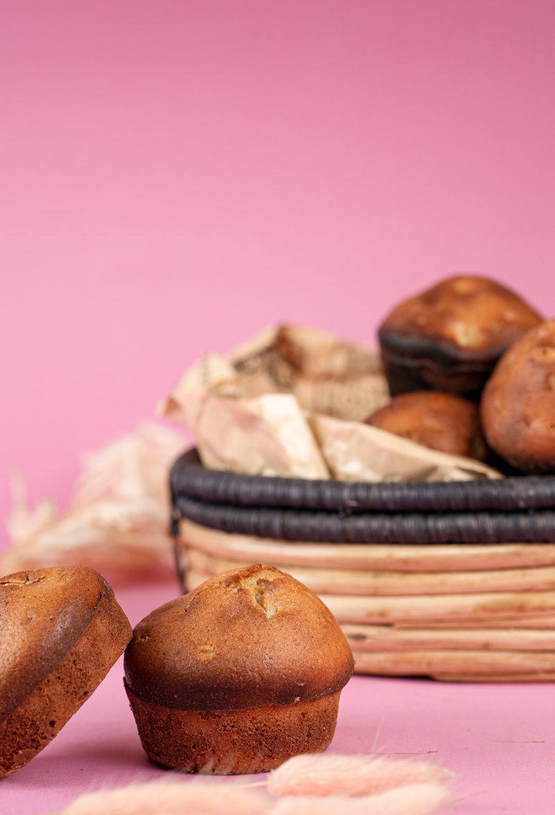 Two cinnamon muffins in front of a basket filled with more muffins on a pink background
