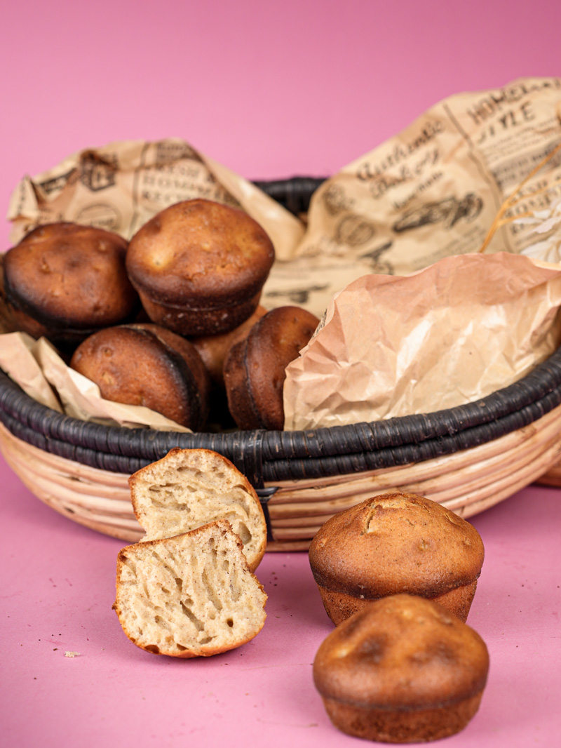Basket of cinnamon muffins with one muffin cut in half to show fluffy texture, on a pink surface